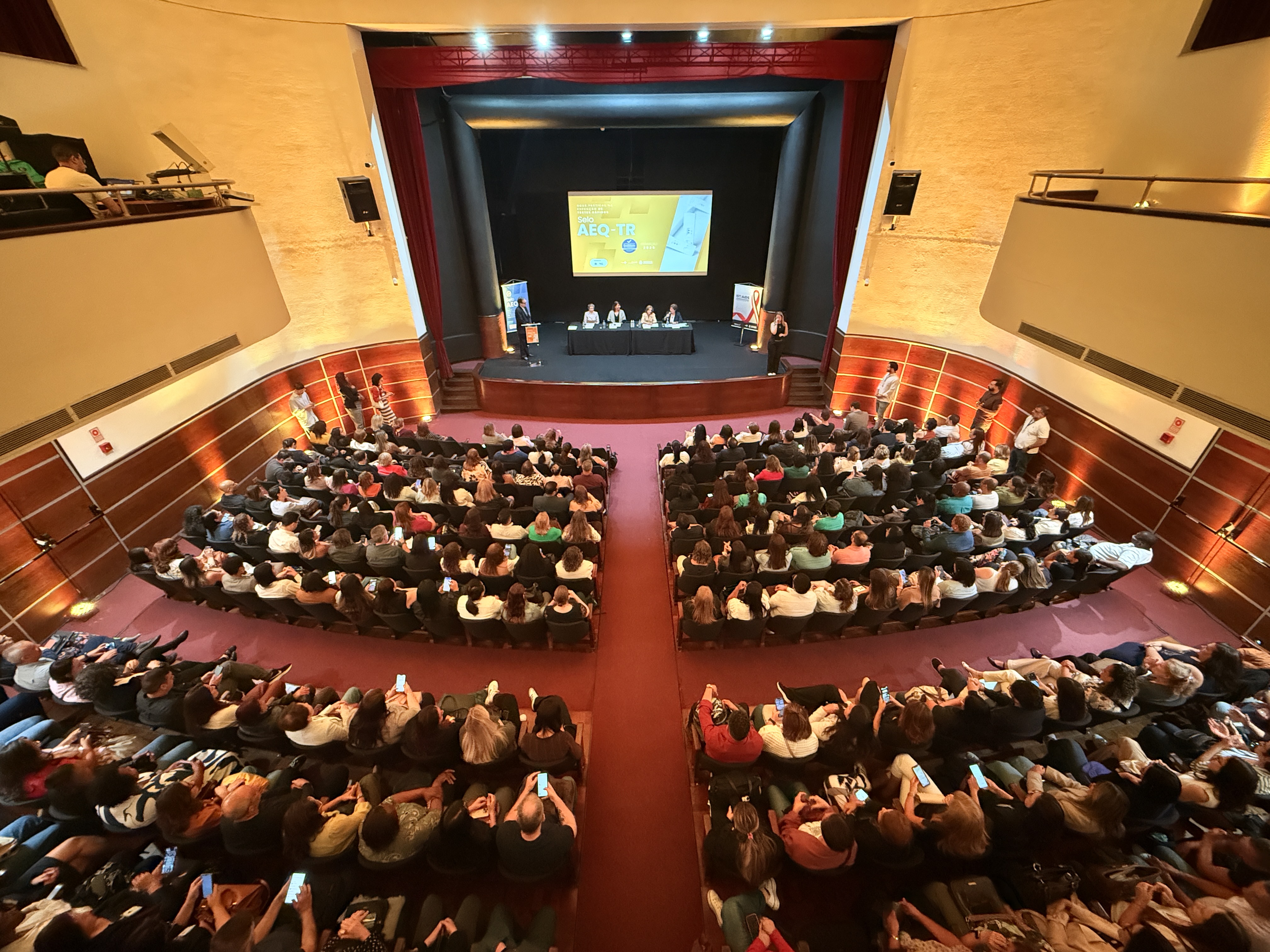 A imagem mostra um auditório lotado, visto de cima, durante um evento. Todas as poltronas estão ocupadas por participantes que assistem a uma mesa composta por quatro pessoas posicionadas no palco. Atrás da mesa, um telão exibe a identidade visual do evento. O espaço é amplo, com paredes laterais curvas e iluminação quente, e algumas pessoas permanecem de pé nas laterais. A disposição da plateia e o enquadramento amplo destacam a grande participação do público.