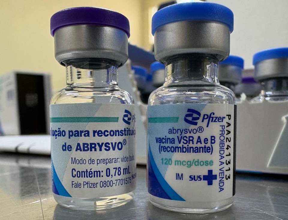A imagem mostra dois frascos de vacina da Pfizer, posicionados lado a lado sobre uma superfície metálica. O frasco à esquerda exibe o rótulo “Solução para reconstituição de ABRYSVO®”, indicando volume de 0,78 mL e instruções de preparo. O frasco à direita traz o rótulo “abrysvo® – vacina VSRA e B (recombinante) – 120 mcg/dose – IM – SUS”, além da indicação “PROIBIDA A VENDA”. Ambos têm tampa roxa e rótulos em tons de azul e branco. Ao fundo, desfocado, é possível ver outros frascos semelhantes organizados em uma bandeja.