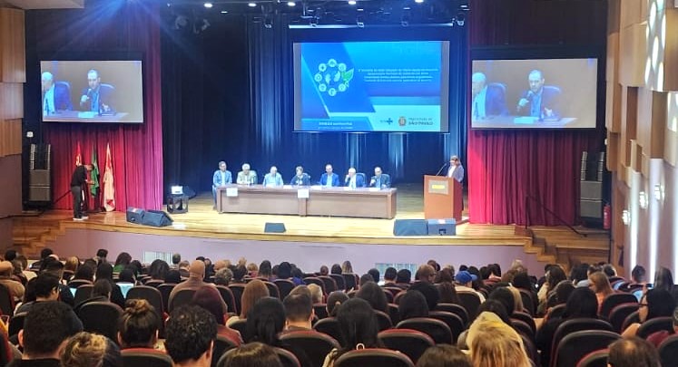 A imagem mostra um auditório lotado de pessoas assistindo a um evento formal. No palco, há uma mesa composta por sete palestrantes sentados, todos de terno, e uma pessoa em pé, atrás de um púlpito, discursando.  Atrás deles, um telão exibe uma apresentação com fundo azul e logotipos — é possível identificar o da Prefeitura de São Paulo. Há também dois telões laterais que projetam a imagem de um dos palestrantes, facilitando a visualização para quem está mais distante.  As cortinas vermelhas laterais emolduram o palco, e o ambiente transmite um clima institucional e acadêmico, provavelmente um seminário ou congresso sobre gestão ou saúde pública.