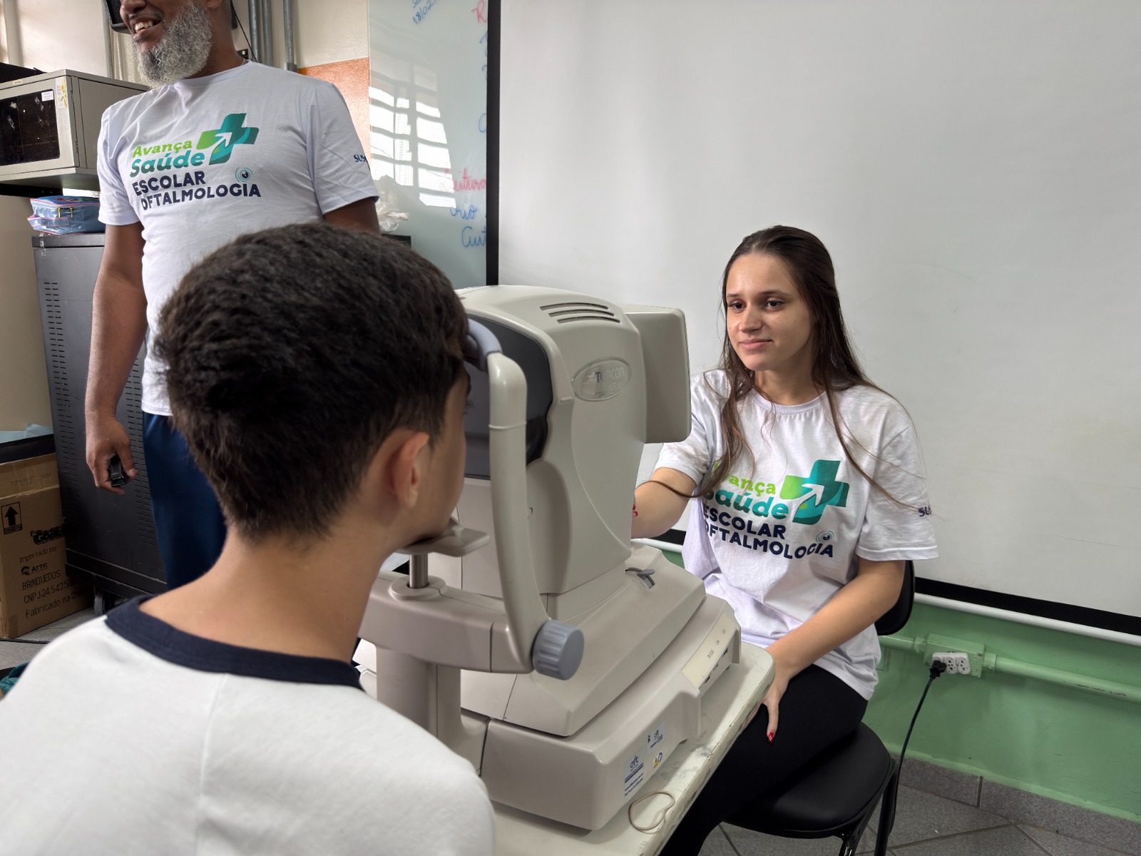 A imagem mostra uma cena de atendimento oftalmológico em ambiente escolar. No centro, uma mulher de pele clara e cabelo castanho longo, solto, veste uma camiseta branca com o logotipo do programa “Avança Saúde Escolar – Oftalmologia”. Ela está sentada diante de um equipamento de exame de vista (autorefrator), operando o aparelho enquanto observa atentamente um estudante. O estudante, visto de costas, tem cabelo curto e castanho-escuro e veste uma camiseta branca com gola azul.