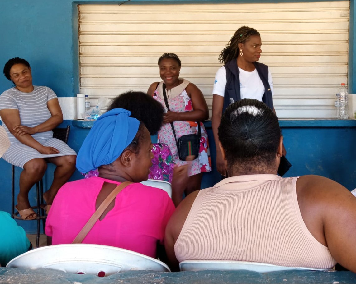A imagem mostra uma cena de encontro comunitário ou atividade social. O ambiente parece ser simples, com paredes azuis e uma porta metálica ao fundo.  No primeiro plano, há mulheres sentadas em cadeiras brancas de plástico, de costas para a câmera. Uma delas veste uma blusa rosa com um lenço azul na cabeça; outra, à direita, usa uma blusa sem mangas em tom claro.  No fundo, estão quatro mulheres em pé ou sentadas:  Uma está de vestido listrado cinza, sentada e sorrindo com os braços cruzados.  Outra, de vestido estampado, sorri e carrega uma bolsinha transversal.  A terceira usa colete azul escuro por cima de uma camiseta branca (possivelmente alguém da área da saúde ou serviço social).  A quarta mulher, parcialmente visível, aparece do lado esquerdo da imagem.  Ao fundo, em cima do balcão azul, há garrafas de água e alguns objetos diversos. A cena transmite um clima de acolhimento, conversa e troca entre as mulheres presentes.