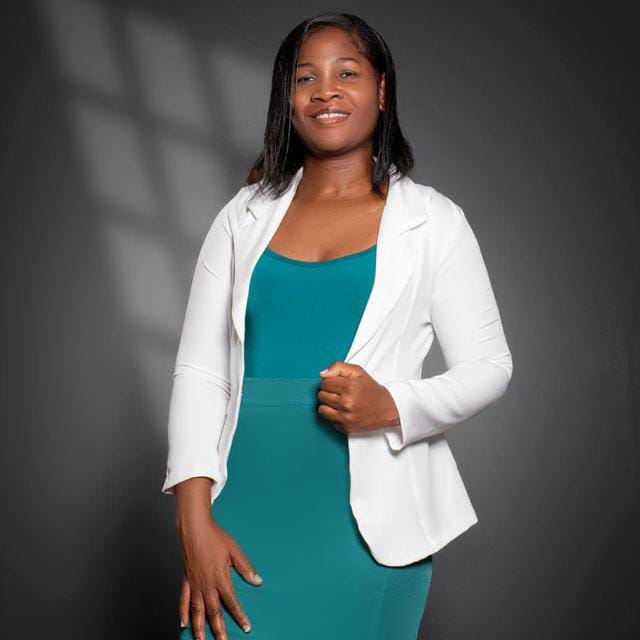 A imagem mostra uma mulher em pé, posando para um retrato em estúdio, com expressão serena e um leve sorriso.  Ela veste um blazer branco sobre um vestido ou conjunto na cor verde-petróleo e segura a lapela do blazer com uma das mãos, enquanto a outra permanece relaxada ao lado do corpo. Seus cabelos são lisos, na altura dos ombros, repartidos de lado, e ela está voltada para a câmera.  O fundo é cinza-escuro, com iluminação suave que cria sombras discretas, destacando a figura da retratada. A composição é simples e elegante, típica de um retrato profissional ou institucional, transmitindo uma imagem de confiança, acolhimento e profissionalismo.