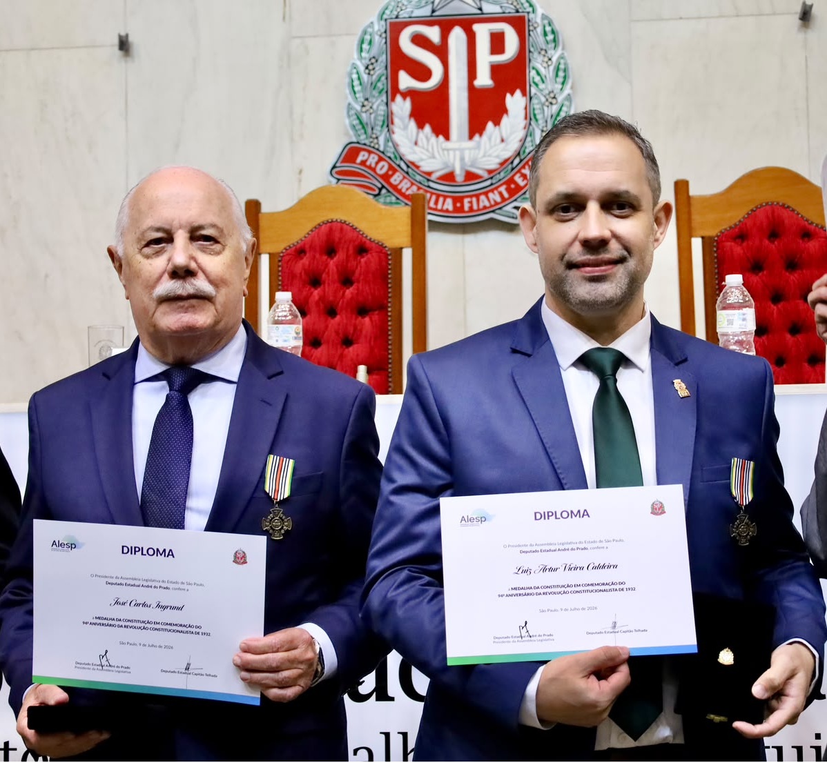 Em um auditório da Assembleia Legislativa do Estado de São Paulo (Alesp), dois homens posam lado a lado em frente ao brasão oficial do Estado de São Paulo e a cadeiras de madeira com estofado vermelho. À esquerda, um homem de cabelos brancos e bigode veste terno azul-marinho, camisa clara e gravata azul, segurando um diploma e a medalha recebida. À direita, Luiz Artur Caldeira, chefe de gabinete da Secretaria Municipal da Saúde de São Paulo, usa terno azul, camisa branca e gravata verde, também segurando o diploma e a medalha da Constituição. Ao fundo, outras autoridades participam da cerimônia, uma delas exibe um diploma. A solenidade ocorreu no plenário da Alesp e homenageou personalidades pelos serviços prestados à sociedade.