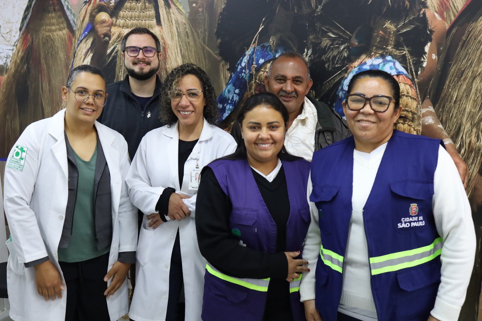 Seis pessoas posam sorrindo em um ambiente interno, diante de um grande painel que mostra imagens de pessoas indígenas usando cocares e vestes tradicionais. No grupo, há três mulheres vestindo jalecos brancos, duas mulheres com coletes azuis da Prefeitura de São Paulo e um homem com jaqueta cinza. Todas e todos olham para a câmera e parecem integrados em um clima de trabalho em equipe e acolhimento.