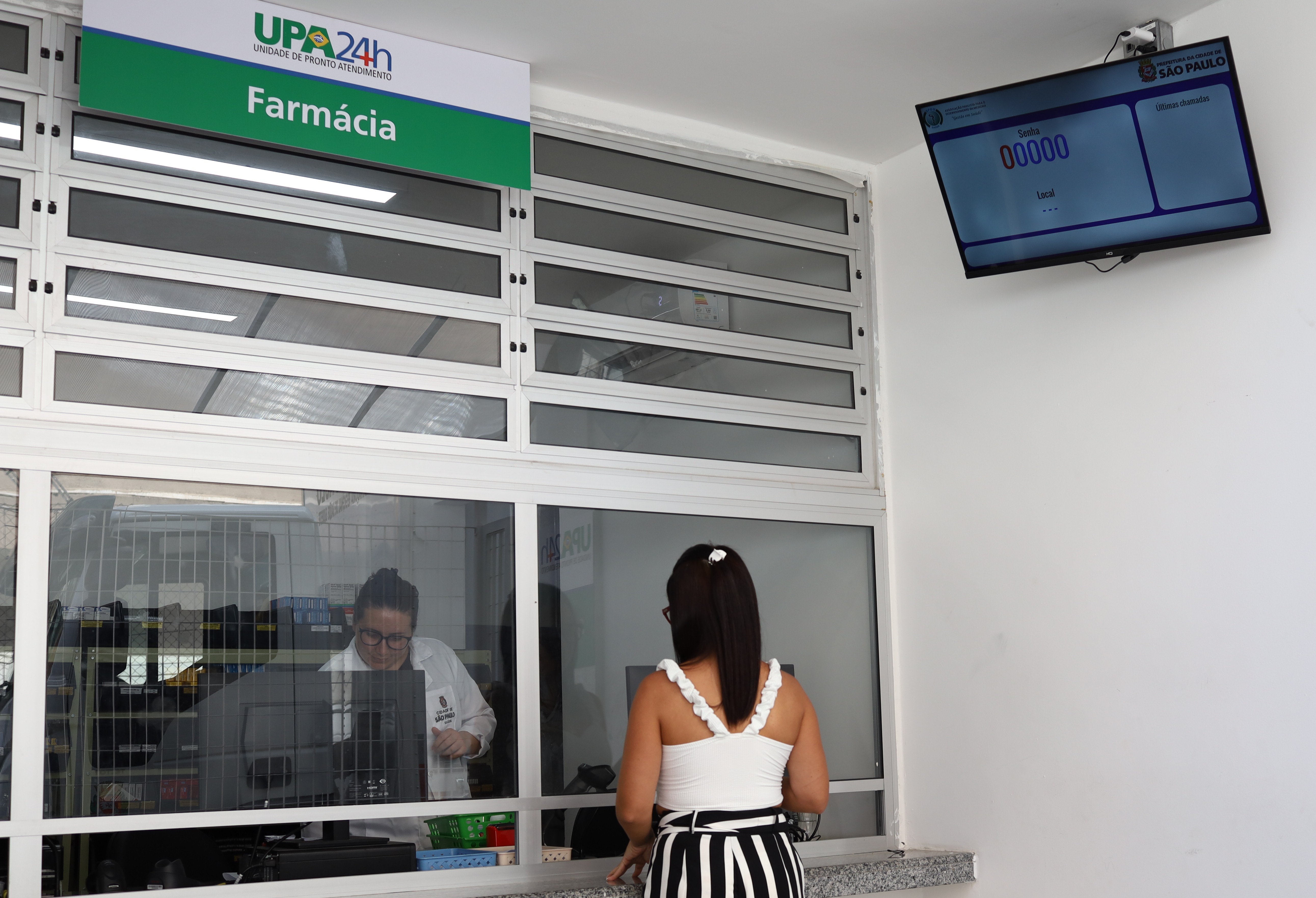 O interior de uma unidade de saúde municipal, especificamente o balcão de atendimento da farmácia. À esquerda, vê-se uma placa indicativa verde e branca com os dizeres