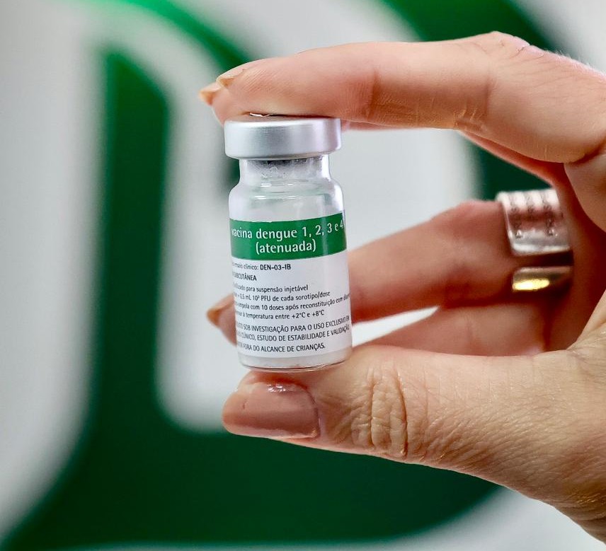 A imagem mostra uma mão segurando um pequeno frasco de vacina. O rótulo indica tratar-se de uma vacina contra dengue (sorotipos 1, 2, 3 e 4), atenuada. O frasco é transparente, com tampa metálica e um rótulo branco e verde contendo informações técnicas.
