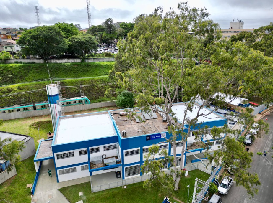 Imagem aérea de um equipamento público de saúde com fachada azul e branca, identificado como Vila Rubi. O prédio é composto por dois blocos térreos e de dois pavimentos, com janelas retangulares e cobertura clara. Ao redor, há áreas gramadas e árvores altas, incluindo uma grande árvore em primeiro plano que parcialmente encobre o edifício. Ao fundo, vê-se um talude verde, uma linha férrea com um trem passando e, mais acima, casas e outros prédios do bairro sob um céu nublado.
