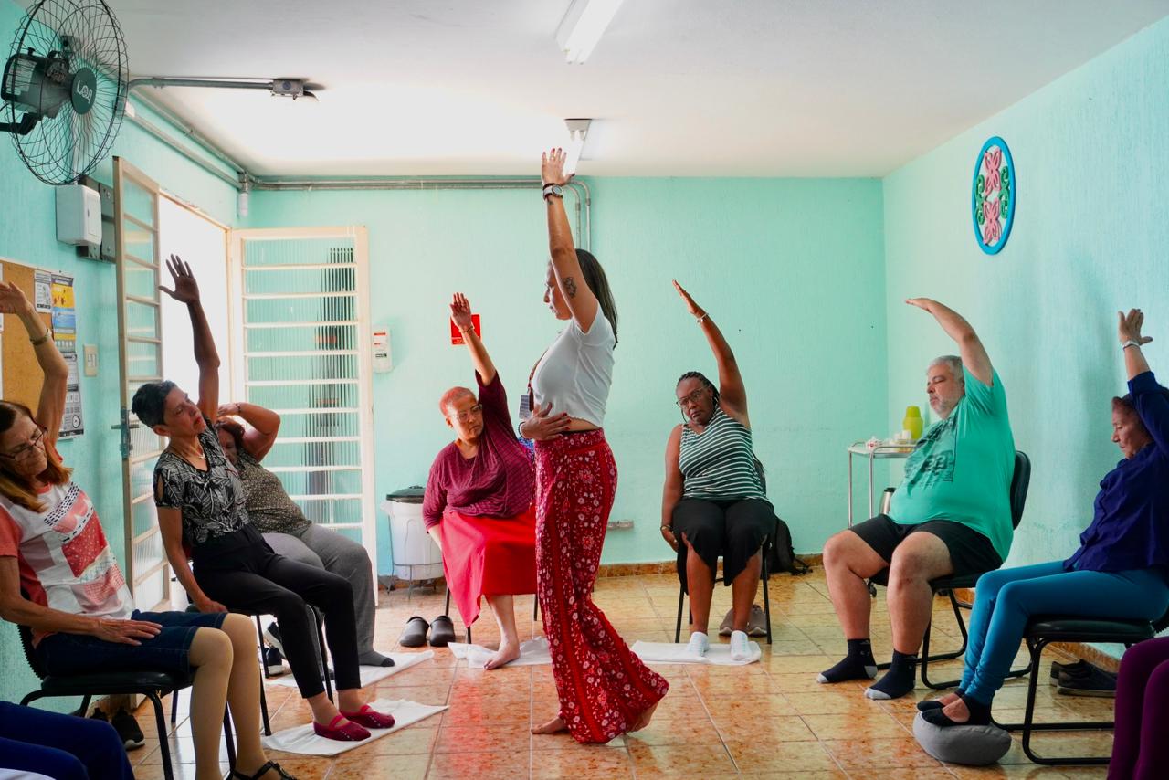 Um grupo de pessoas sentadas, a maioria mulher, está com o braço esquerdo levantado. São conduzidas pela professora que está ao centro. Ela também está com o braço levantado, mas em pé. Ela veste uma camiseta branca e uma calça vermelha estampada 