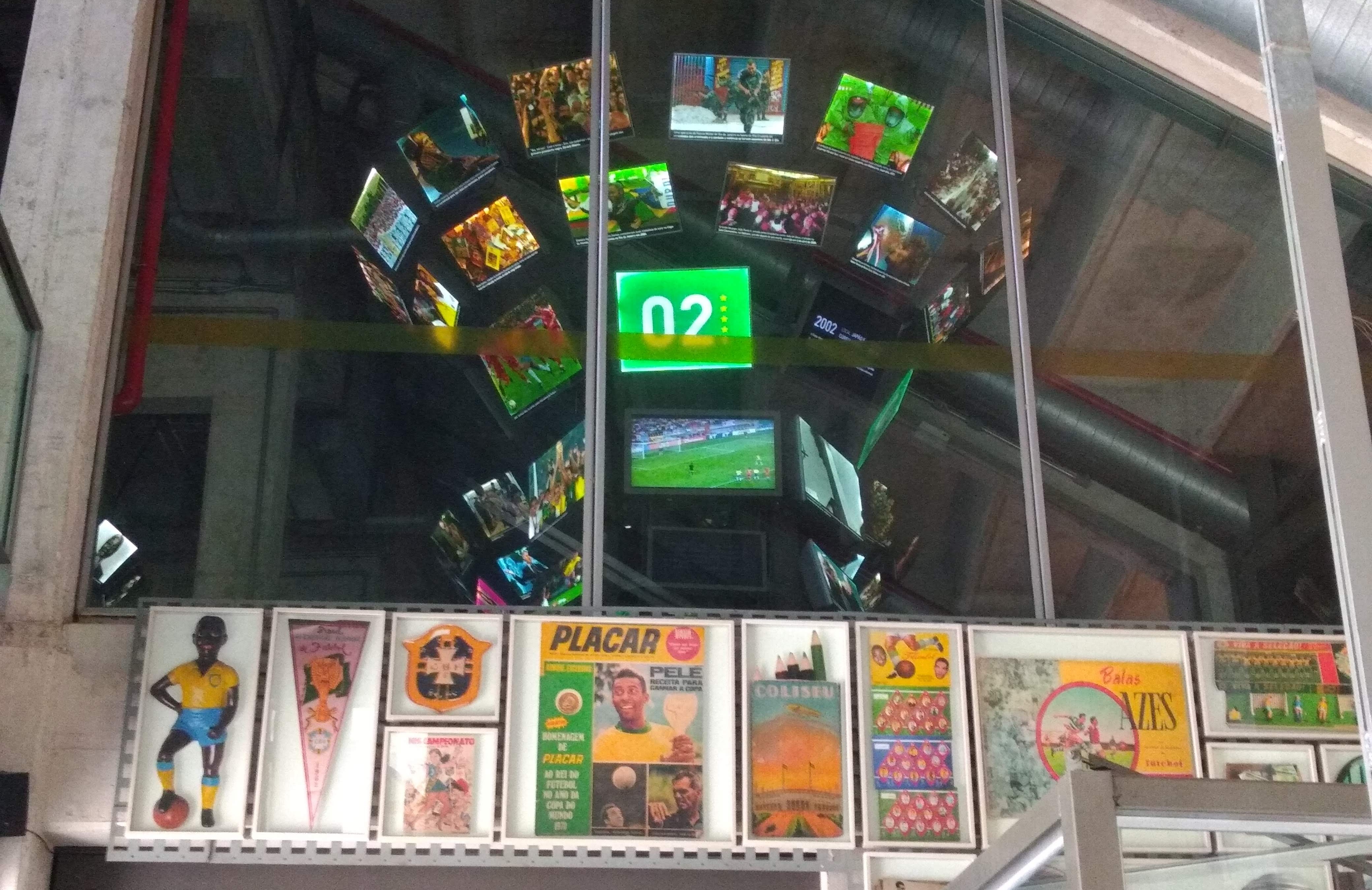 A imagem mostra uma parede com diversos quadros e pôsteres relacionados ao futebol, incluindo revistas, escudos e ilustrações históricas. Acima dos quadros, há uma grande divisória de vidro onde se refletem várias telas de televisão formando um grande painel circular, exibindo imagens de partidas de futebol e outros eventos esportivos. No centro superior, destaca-se um sinal luminoso verde com o número “02”. O ambiente remete a um espaço cultural ou museu esportivo, valorizando a memória do futebol brasileiro.