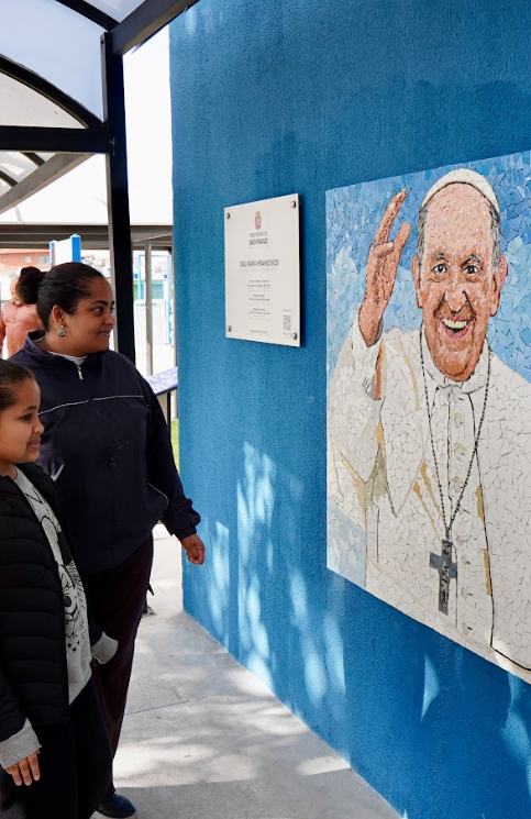 Na imagem, duas pessoas observam atentamente um mural em uma parede azul. O mural retrata uma figura religiosa vestida com indumentária branca, um crucifixo pendurado no pescoço e uma das mãos erguidas em gesto de saudação ou bênção. Ao lado do mural, há uma placa com inscrições, fixada na mesma parede. Um ambiente coberto proporciona sombra parcial na área. A cena transmite um momento de contemplação e respeito diante da representação religiosa.