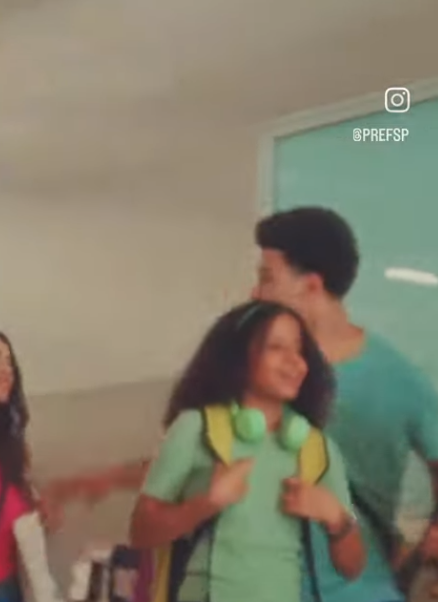 Cena em ambiente interno com três adolescentes caminhando juntos, sorrindo. À frente, uma menina com cabelo crespo solto, usando uma camiseta verde clara e mochila amarela nas costas, com um fone de ouvido verde pendurado no pescoço. Ao lado dela, à esquerda, aparece parcialmente outra menina, de cabelos compridos e escuros, vestindo rosa. À direita, um menino de camiseta azul-clara, de perfil, também sorri enquanto anda ao lado das colegas. Ao fundo, uma parede clara com uma grande janela de vidro. No canto superior direito da imagem, há o logo do Instagram e o nome de usuário @PREFSP. O ambiente transmite uma atmosfera alegre e descontraída, possivelmente em um contexto escolar.