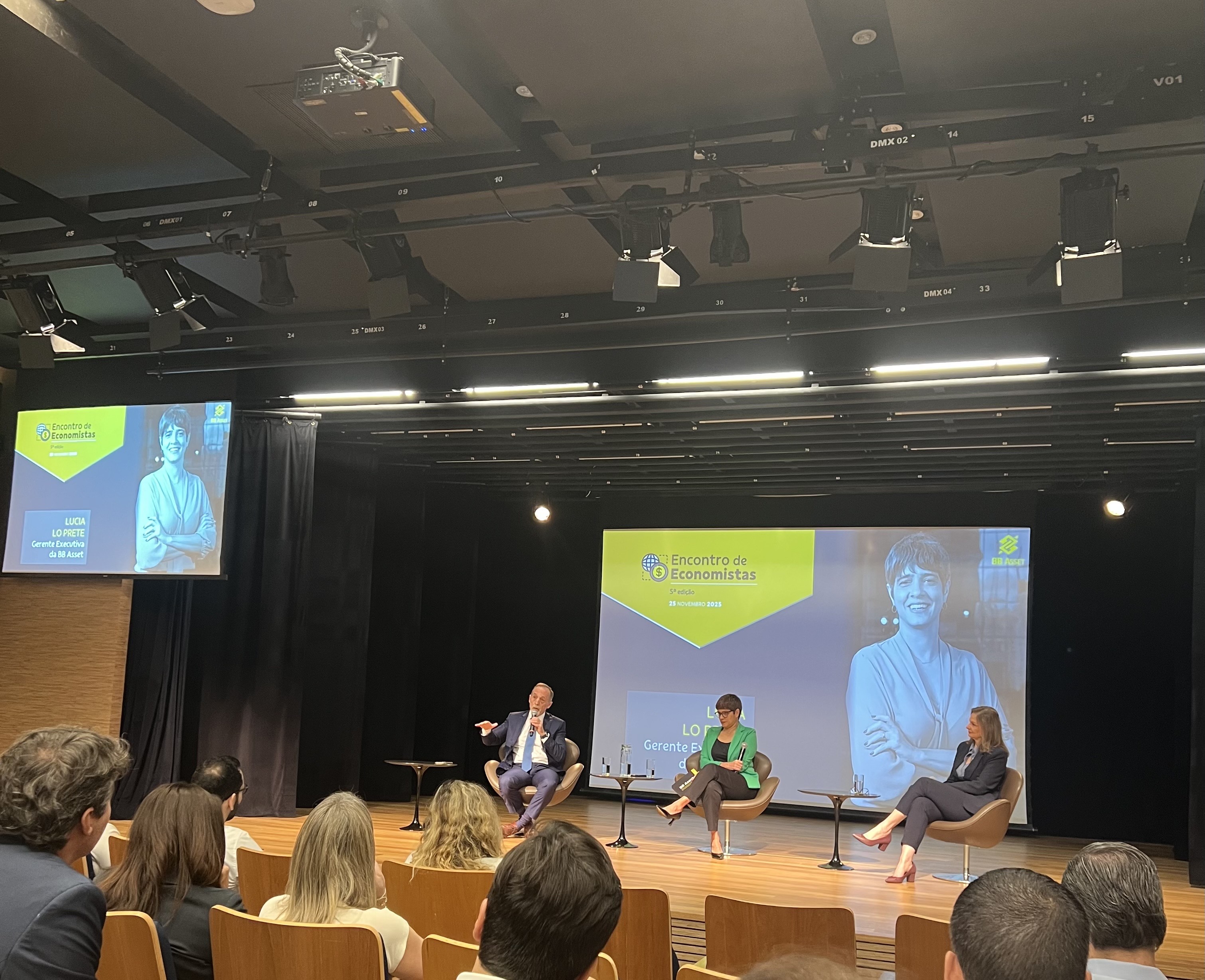A imagem mostra um auditório corporativo com pessoas sentadas em cadeiras de madeira, assistindo a um painel no palco. Três palestrantes estão sentados em poltronas separadas, cada um com um pequeno mesa lateral e microfone. O palco é iluminado por luzes de teto e há cortinas pretas ao fundo. Atrás dos palestrantes há uma grande tela projetando o título do evento: “Encontro de Economistas – 3ª edição – 29 de novembro de 2023”. A tela exibe também uma fotografia de uma pessoa sorridente e o nome “Lúcia Lepeltier – Gerente Executiva do BB Asset”, conforme aparece no slide. À esquerda do palco há outro telão menor mostrando a mesma imagem e texto.