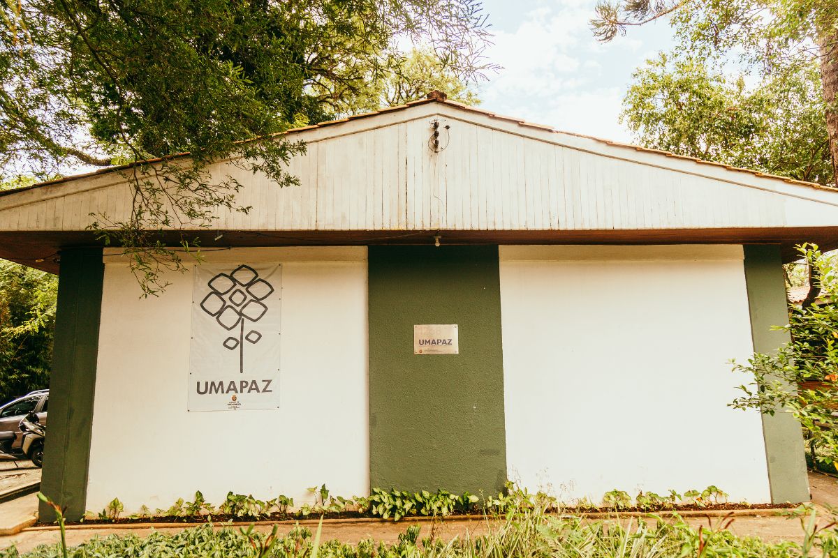 A imagem mostra a fachada de um prédio simples, cercado por vegetação. Na parede, há uma placa e um banner com a logomarca da UMAPAZ (Universidade Aberta do Meio Ambiente e Cultura de Paz), uma instituição voltada para educação ambiental e sustentabilidade, localizada no Parque Ibirapuera, em São Paulo. O edifício apresenta tons de verde e branco, integrando-se à paisagem natural ao redor.