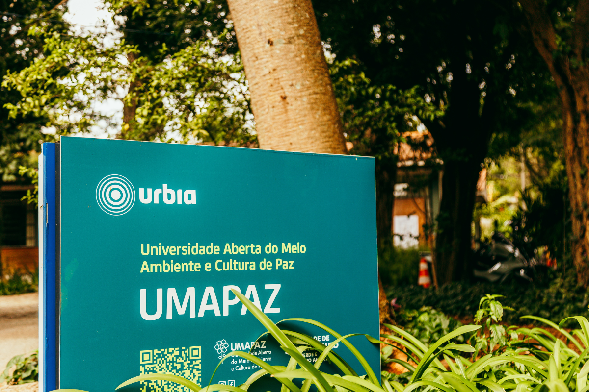 Foto de uma placa verde com o logotipo da Urbia e os dizeres “Universidade Aberta do Meio Ambiente e Cultura de Paz – UMAPAZ”. A placa está posicionada em um ambiente arborizado, com plantas em primeiro plano e árvores ao fundo, indicando que o local está inserido em um espaço natural. Também é possível ver parte de uma construção e um cone laranja ao fundo.