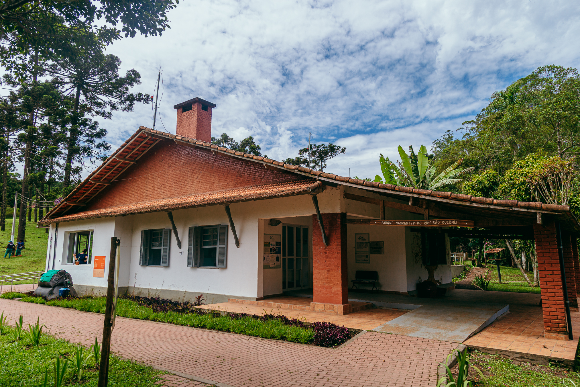 A foto mostra um prédio branco da Escola de Agroecologia de Parelheiros, com telhado de telhas vermelhas. Há uma chaminé de tijolos no telhado. O prédio tem janelas e portas. Uma área coberta com pilares de tijolos está à direita. Um caminho de tijolos leva ao prédio. À esquerda, há árvores altas e verdes. Uma pessoa está sentada no chão perto das árvores. O céu tem nuvens brancas e manchas de azul.
