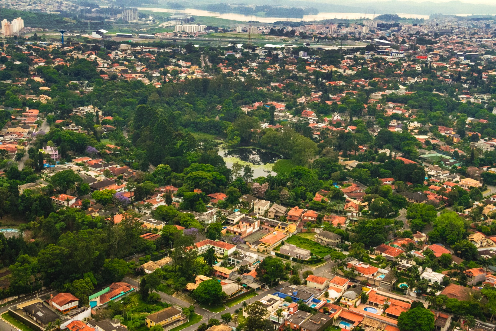 A imagem mostra uma vista aérea de um bairro arborizado com muitas casas de telhado vermelho e ruas bem distribuídas. No centro da imagem, há uma área com bastante vegetação e um lago cercado por árvores. Ao fundo, observa-se uma região mais urbanizada, com prédios e vias movimentadas, além de um rio ou represa. A paisagem apresenta um equilíbrio entre natureza e urbanização, destacando uma área residencial de alto padrão.