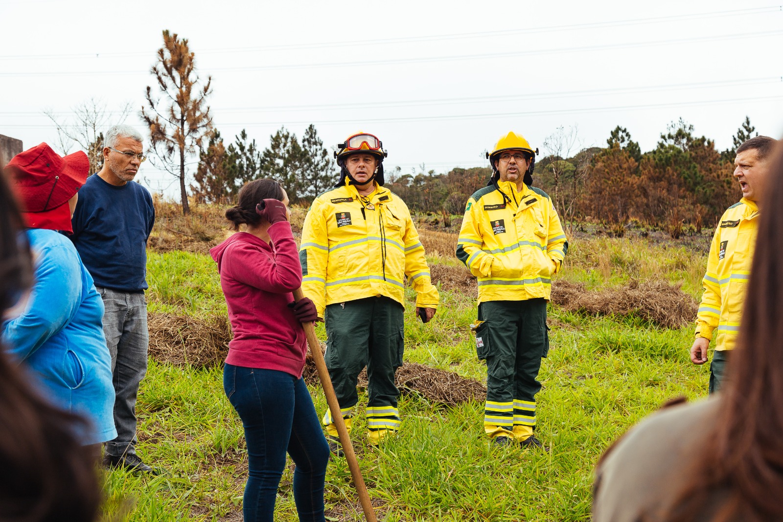 A imagem mostra dois brigadistas da Operação Fogo Zero em um treinamento para a população para casos de incêndio em áreas verdes. Os bombeiros estão de trajes amarelo segurando um enxada