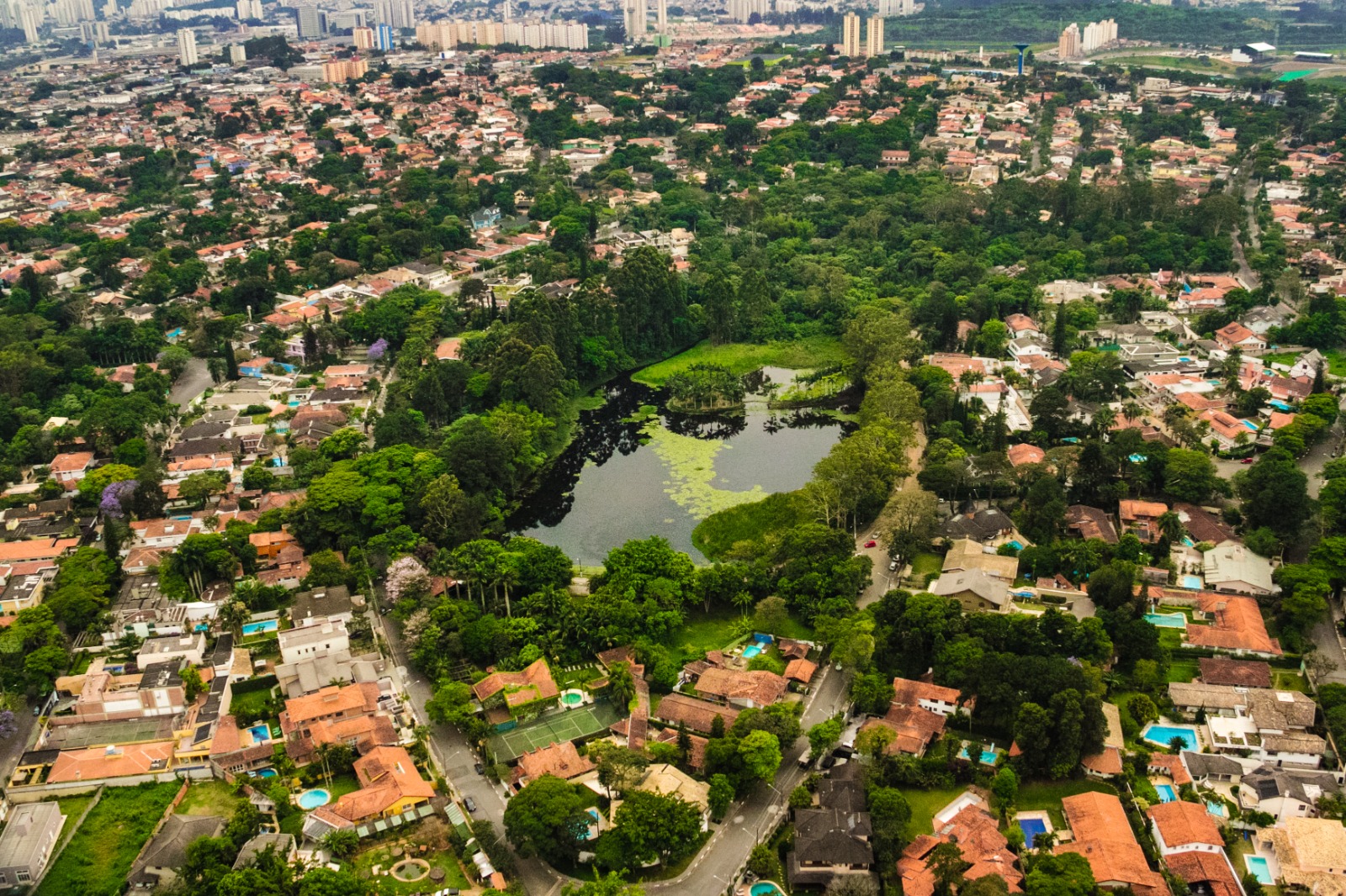 Esta imagem mostra uma vista aérea de uma área urbana densamente arborizada, com um grande corpo d’água cercado por vegetação e residências. O cenário mistura espaços naturais preservados com construções, incluindo casas de alto padrão com piscinas.