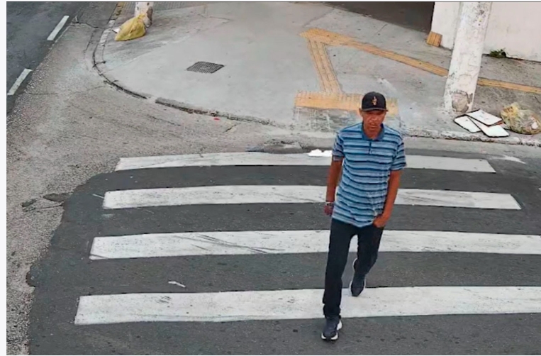 Homem de blusa azul listrada,calça escura e boné preto, atravessando a via.