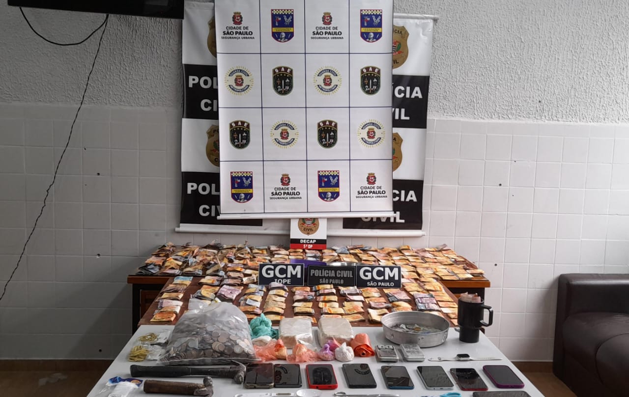 Foto mostra uma mesa uma grande quantia de dinheiro, drogas, armas e celulares que forma apreendidos pela Guarda Civil Metropolitana. Ao fundo um cartaz com o logo da cidade de São Paulo e da Guarda Civil Metropolitana