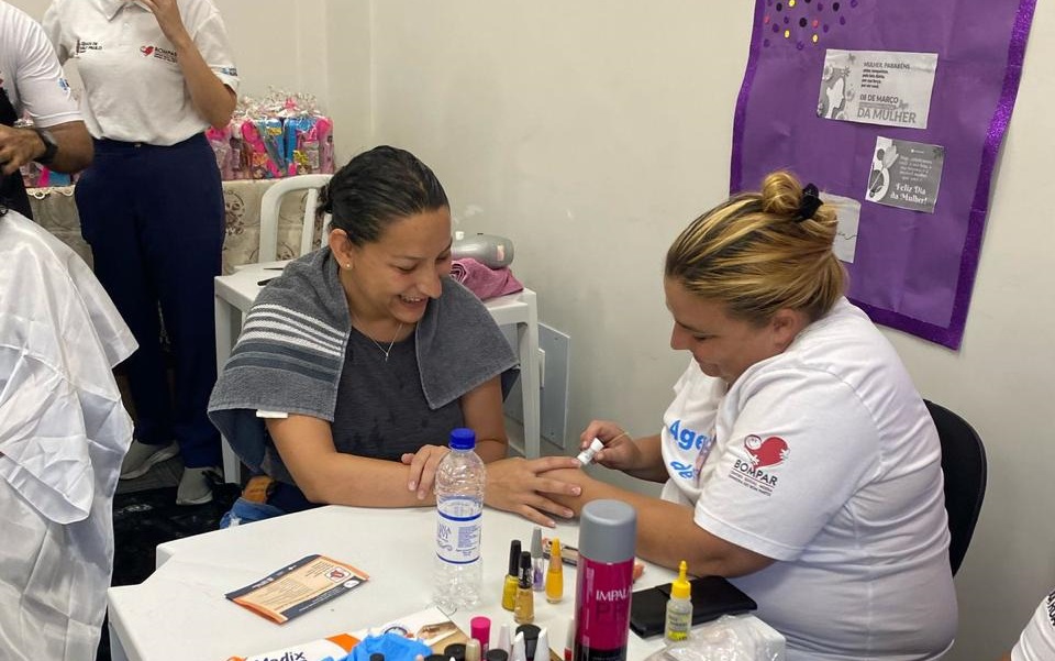 Em um ambiente decorado para o Dia da Mulher, uma profissional, vestindo uma camisa branca com o logotipo do projeto, realiza o serviço de manicure em uma mulher. A cliente está sentada à mesa, com uma toalha nos ombros, enquanto a manicure pinta suas unhas. Na mesa, há diversos frascos de esmalte e produtos de beleza. Ao fundo, um mural roxo com informações sobre a data comemorativa e outras pessoas participando das atividades do evento.