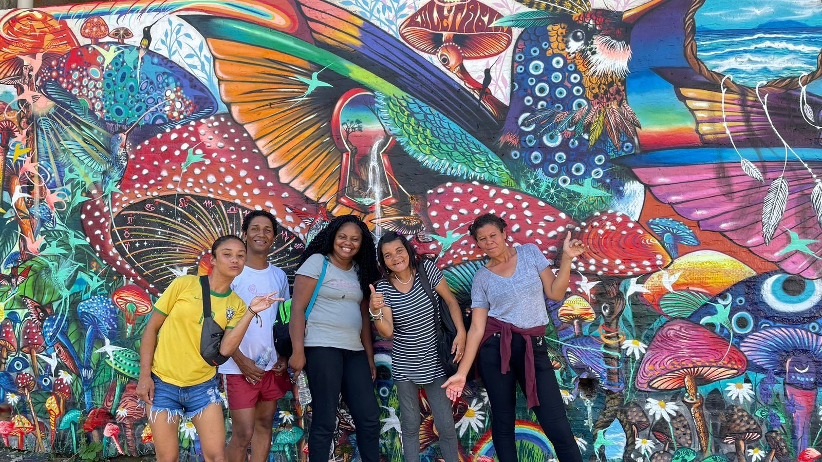 Imagem de um grupo de cinco mulheres lado a lado posando para uma foto. Elas estão em frente a um muro colorido e personalizado com vários desenhos de cogumelos