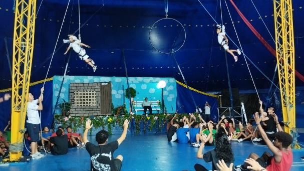 Imagem da parte interna de um Circo Social. Na imagem há diversas crianças e adolescentes sentadas no chão em roda assistindo uma apresentação artística. No fundo da imagem e na parte superior há uma lona azul escura.