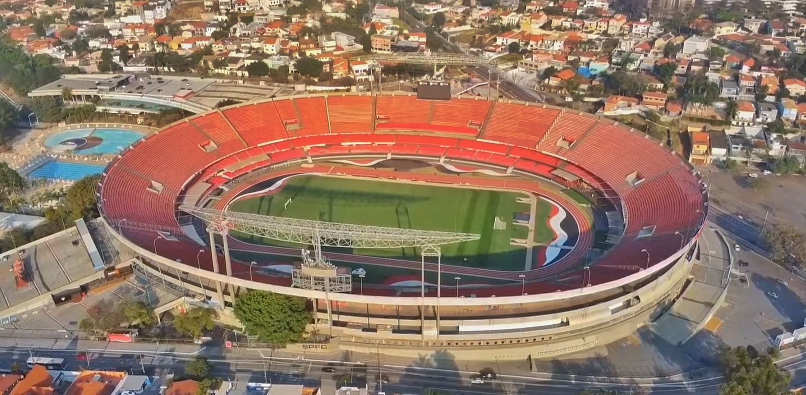 Vista aérea de um estádio desportivo com arquibancadas vermelhas e campo verde. A composição da imagem é de uma vista aérea de um grande estádio desportivo. O estádio é circular, com arquibancadas vermelhas que circundam todo o campo verde. Há estruturas metálicas de suporte que se projetam para cima do estádio. A estrutura se sobrepõe a uma pequena parte de uma cidade com edifícios, ruas e árvores. A imagem mostra a grandiosidade da estrutura do estádio e seu tamanho em relação à paisagem circundante. O tempo parece ser um dia claro. Os principais assuntos da imagem são o estádio e o campo. O estádio apresenta arquibancadas vermelhas em forma de anel, com degraus numerosos. O campo de jogo é de cor verde grama e parece estar vazio. A presença de uma pista ao redor do campo é visível. A estrutura geral do estádio está em destaque, revelando suas características arquitetônicas de forma nítida. A imagem é feita por meio de uma fotografia aérea ou tomada de uma perspectiva de drone. Os detalhes da estru