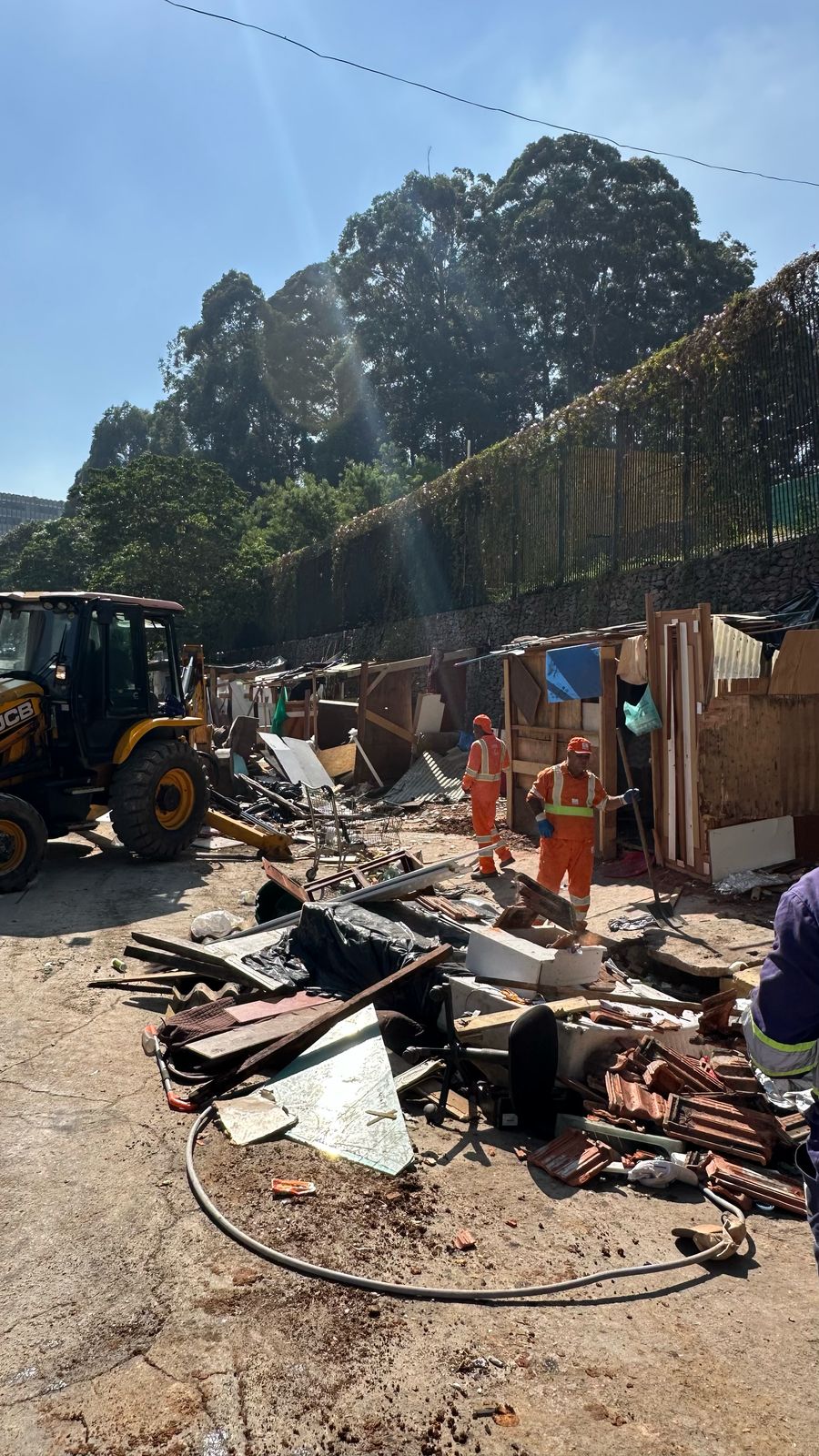 Imagem: Uma foto tirada durante o dia, mostrando uma área ao ar livre que parece ter sido um acampamento ou uma favela em processo de demolição. A luz do sol está forte, indicando um dia claro e ensolarado.  Elementos principais:  Primeiro plano: Uma grande quantidade de entulho e detritos espalhados pelo chão de concreto. Há pedaços de madeira, telhas, objetos metálicos retorcidos e outros materiais de construção quebrados. Um longo tubo preto de borracha está enrolado no chão. Meio do plano: Dois homens vestindo uniformes laranja com listras reflexivas estão no centro da imagem. Eles parecem estar trabalhando na limpeza ou demolição do local. Um deles está de costas para a câmera, enquanto o outro está de perfil. Fundo: Uma série de estruturas improvisadas, como barracos feitos de madeira e outros materiais, são visíveis. Essas estruturas estão parcialmente demolidas, com partes faltando ou quebradas. Ao fundo, há uma cerca alta de metal e árvores frondosas, sugerindo que o local pode estar próximo a uma área arborizada ou parque. Lateral esquerda: Uma retroescavadeira amarela e preta da marca JCB é parcialmente visível, indicando que maquinário pesado está sendo usado na operação. Lateral direita: Uma pequena parte de uma pessoa vestindo um uniforme azul é visível, sugerindo que há mais pessoas envolvidas na operação fora do quadro. Cores: As cores predominantes na imagem são o marrom e o cinza do entulho, o laranja dos uniformes dos trabalhadores e o verde das árvores ao fundo. O céu está claro e azul, indicando um dia ensolarado.  Atmosfera: A imagem transmite uma sensação de desordem e destruição, mas também de trabalho e atividade, com os trabalhadores limpando a área. A presença da retroescavadeira sugere que a demolição ou limpeza está em andamento.  Informações adicionais: A imagem foi tirada na vertical e tem uma boa iluminação, permitindo que os detalhes sejam claramente visíveis. A qualidade da foto é boa, com cores vibrantes e foco nítido.