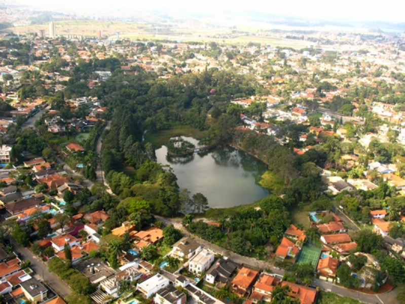 A foto mostra uma vista aérea e distante de um bairro chamado Interlagos, em São Paulo. No centro da imagem, há um lago conhecido como Parque do Laguinho cercado por árvores, e ao redor, é possível ver um grande número de casas com telhados de cor laranja e marrom.