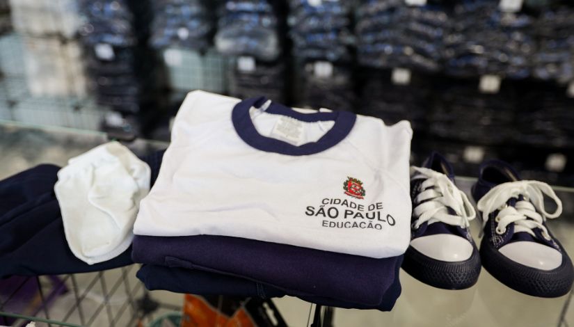 A imagem mostra um conjunto de uniforme escolar organizado sobre uma superfície, possivelmente uma mesa ou carrinho. No centro, há uma camiseta branca dobrada, com gola azul-escura. No lado esquerdo do peito da camiseta, aparece um brasão colorido e o texto “Cidade de São Paulo – Educação”, indicando que é um uniforme da rede municipal de ensino.  Abaixo da camiseta, vê-se uma peça de roupa azul-escura dobrada, provavelmente uma calça ou bermuda do uniforme. À esquerda do conjunto, há um par de meias brancas dobradas. À direita, aparece um par de tênis infantis nas cores azul-marinho e branco, com cadarços brancos e biqueiras claras.