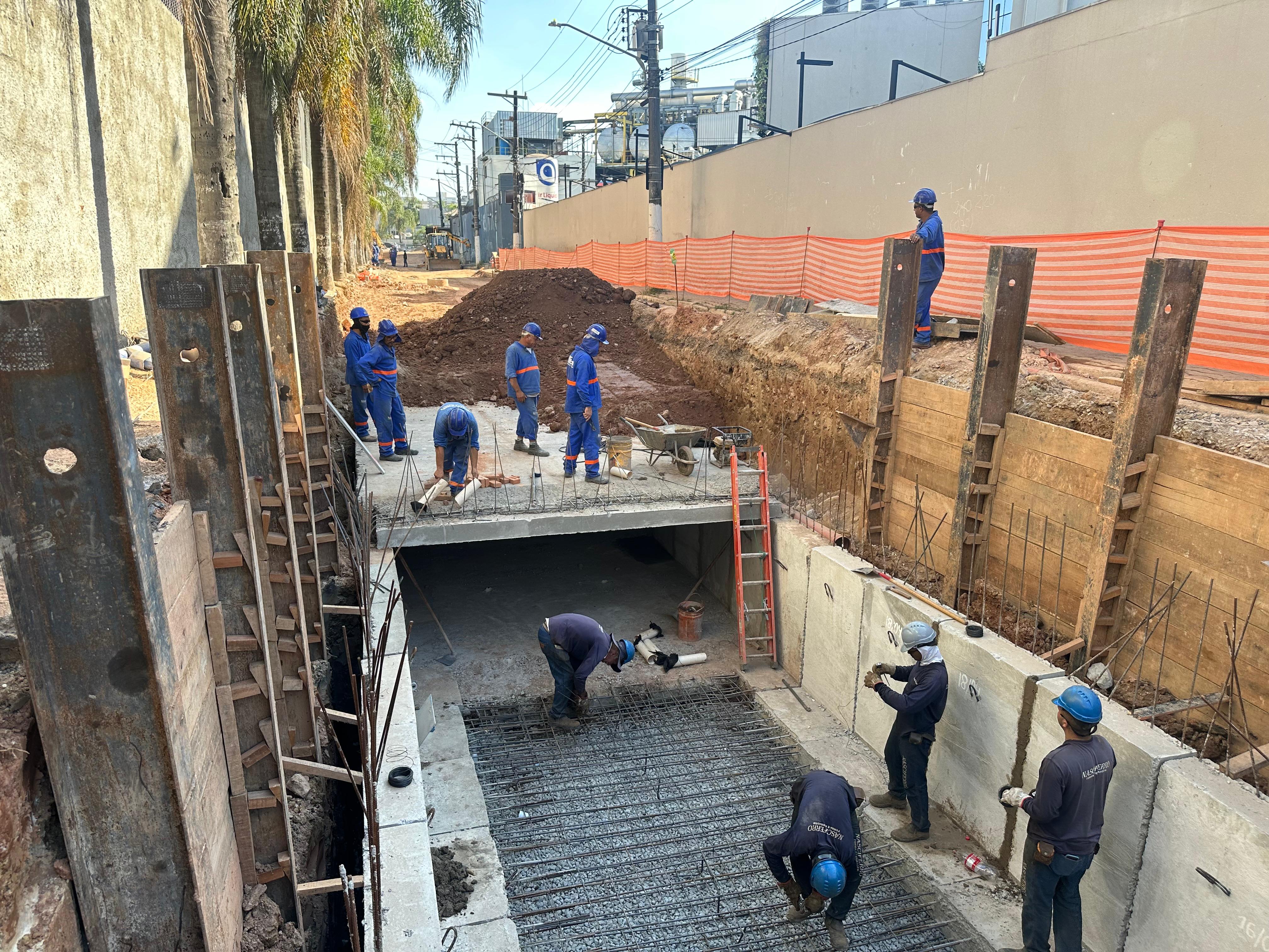 Grupo de trabalhadores trabalhando na construção de uma galeria. Nas laterais a imagem mostra perfis de metal e paredes de concreto. No piso da galeria montam uma armação de ferro para concretagem posterior.