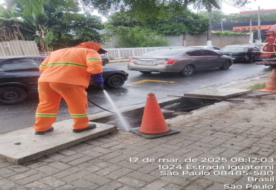 Na Estrada do Iguatemi o zelador vestindo uniforme na cor laranja, empunha uma mangueira com água e faz a lavagem de dois bueiros que encontram-se abertos e sinalizados com dois cones. Logo abaixo encontra-se a programação, em planilha separada por linhas em azul.