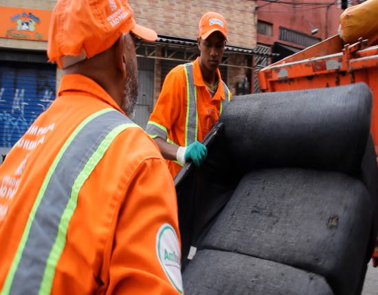 dois zeladores em uma rua pública usando uniformes e bonés na cor laranja e luvas verdes realizam a operação cata bagulho e estão recolhendo um sofá direcionando o mesmo para o caminhao da empresa estacionado próximo