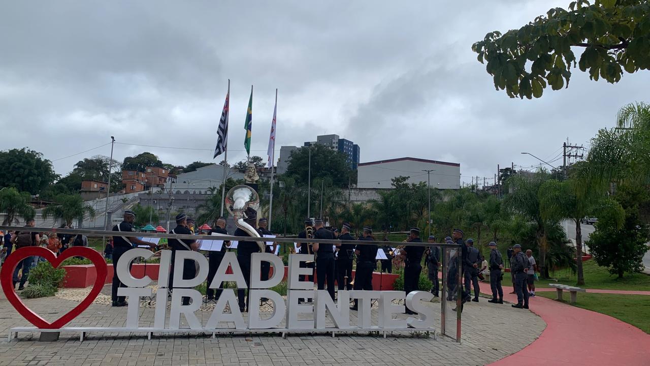 à frente da imagem o nome de Cidade Tiradentes com o coração indica o amor ao bairro, logo  atrás a banda da GCM posicionada com instrumentos musicais e ao fundo as bandeiras hasteadas