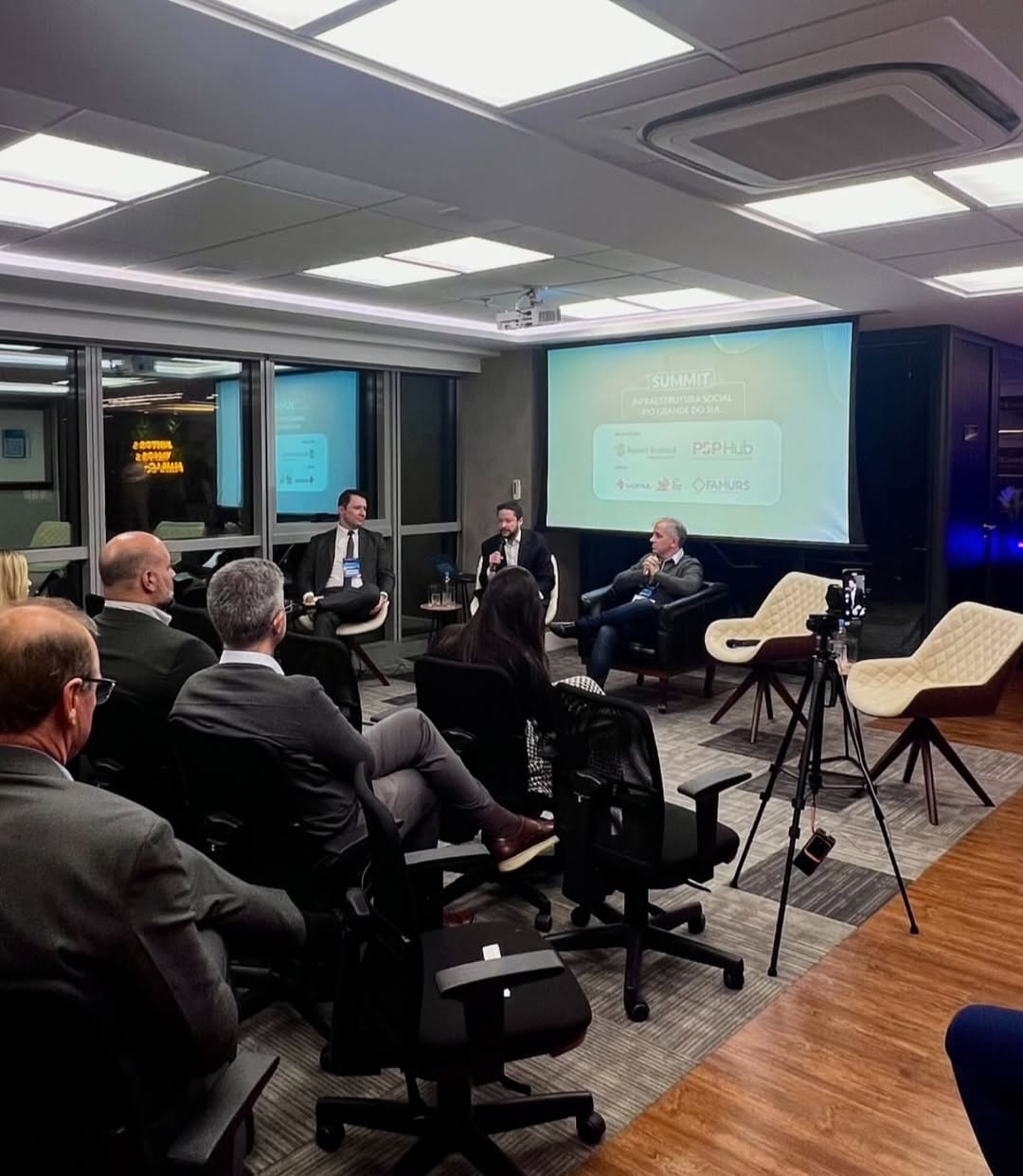 Foto do Presidente da SPDA, Hélio Rubens de Oliveira Mendes, palestrando em evento, sentado com mais dois palestrantes, sendo ouvidos por um grupo de pessoas também sentadas, em sala fechada, com disposição espacial de congresso, com projeção de apresentação atrás dos palestrantes.