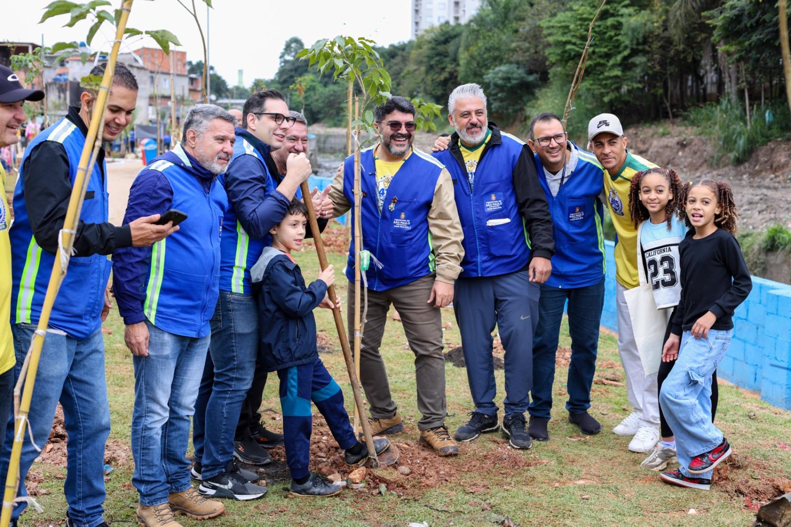 Parte da equipe da Subprefeitura Itaim Paulista, com coletes azuis, posa com três munícipes — duas crianças e um adulto — durante o plantio de uma das 415 árvores em celebração aos 415 anos do Itaim Paulista.