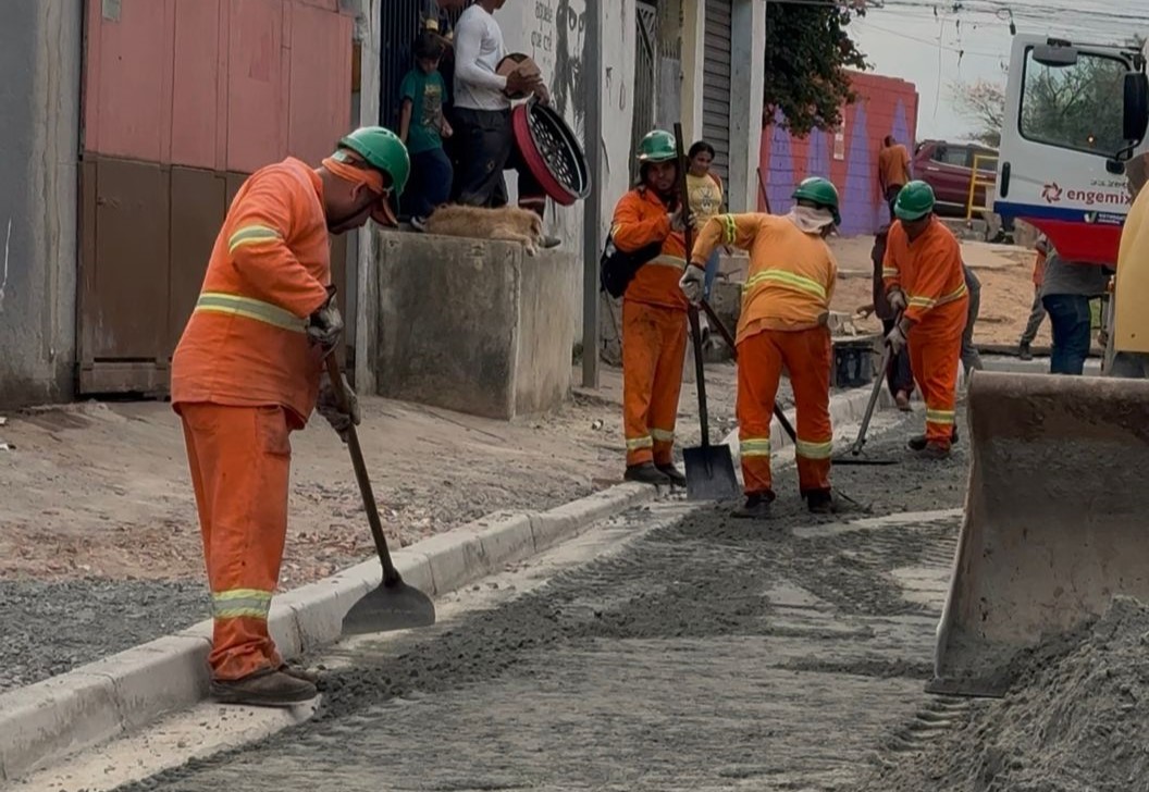 Quatro trabalhadores, usando uniformes laranjas e capacetes verdes, realizam a pavimentação da Rua Ita Rio de Janeiro.