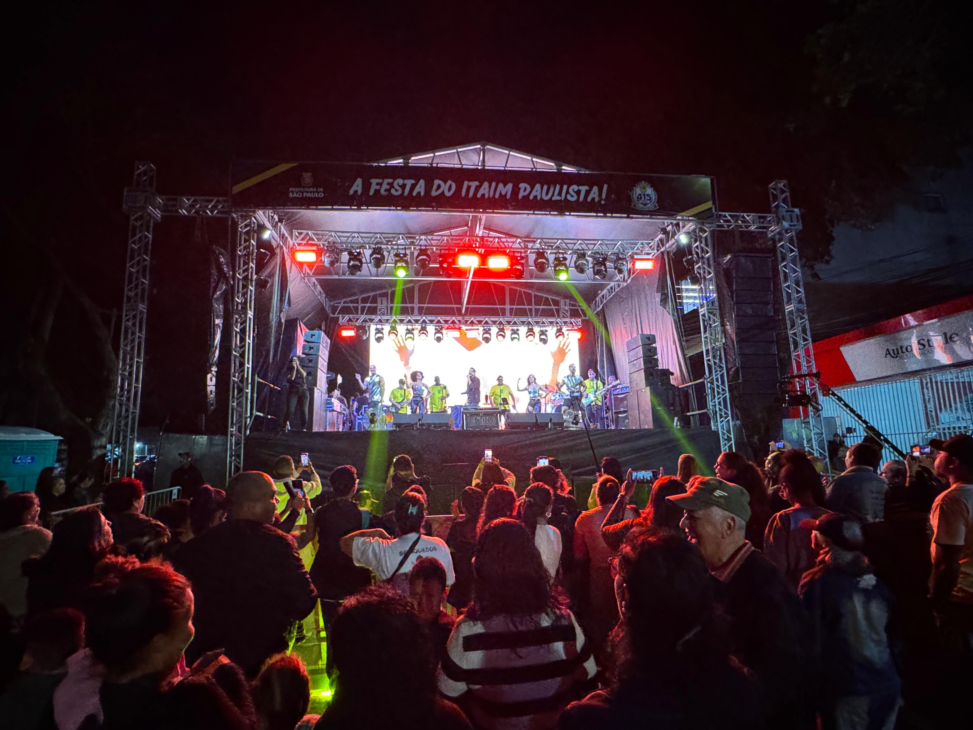 Palco da festa de 415 anos do Itaim Paulista, com a inscrição “A Festa do Itaim Paulista” e os logos da Prefeitura de São Paulo e da comemoração. O cantor Latino se apresenta diante do público.