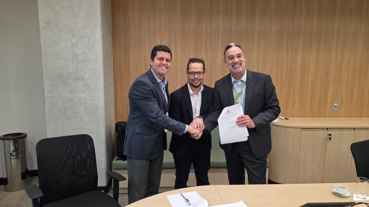 Três homens em pé, sorrindo e apertando as mãos em uma sala de reuniões. Um deles segura documentos assinados. Ao fundo, parede de madeira clara e mesa de reuniões com papéis, xícara e copo com água sobre ela.
