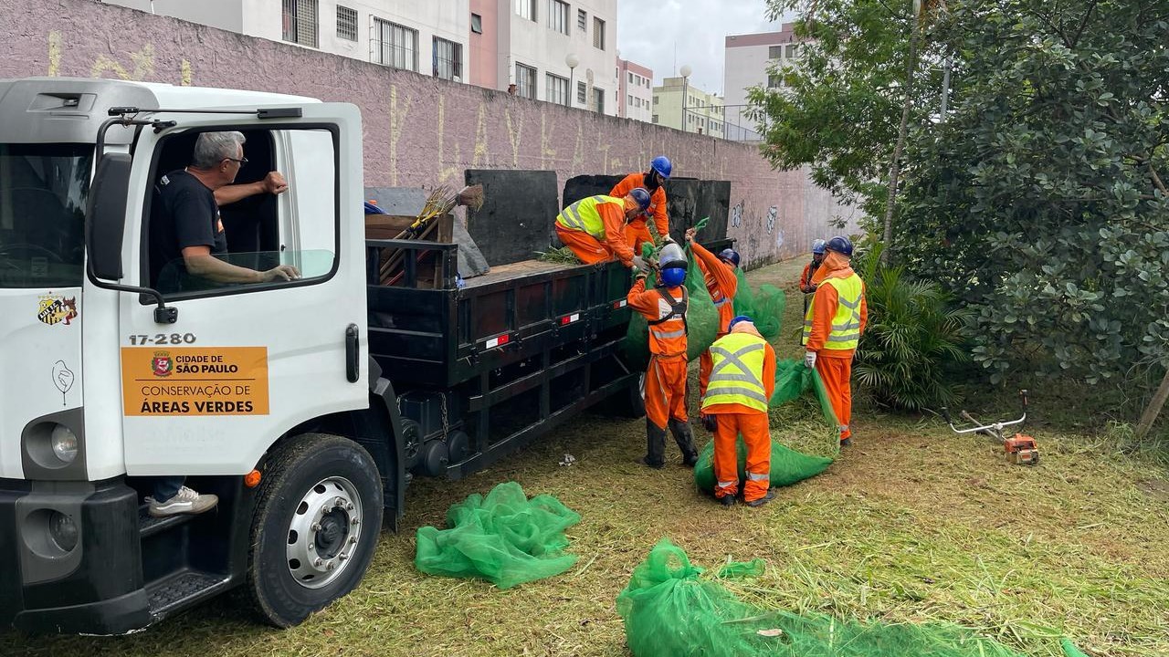 #PraCegoVer: Imagem mostra seis funcionários da Prefeitura de São Paulo recolhendo sacos plásticos cheios de mato cortado e colocando-os em um caminhão. Eles estão utilizando EPIs (Equipamentos de Proteção Individual), uniformes de cor laranja e coletes amarelos.