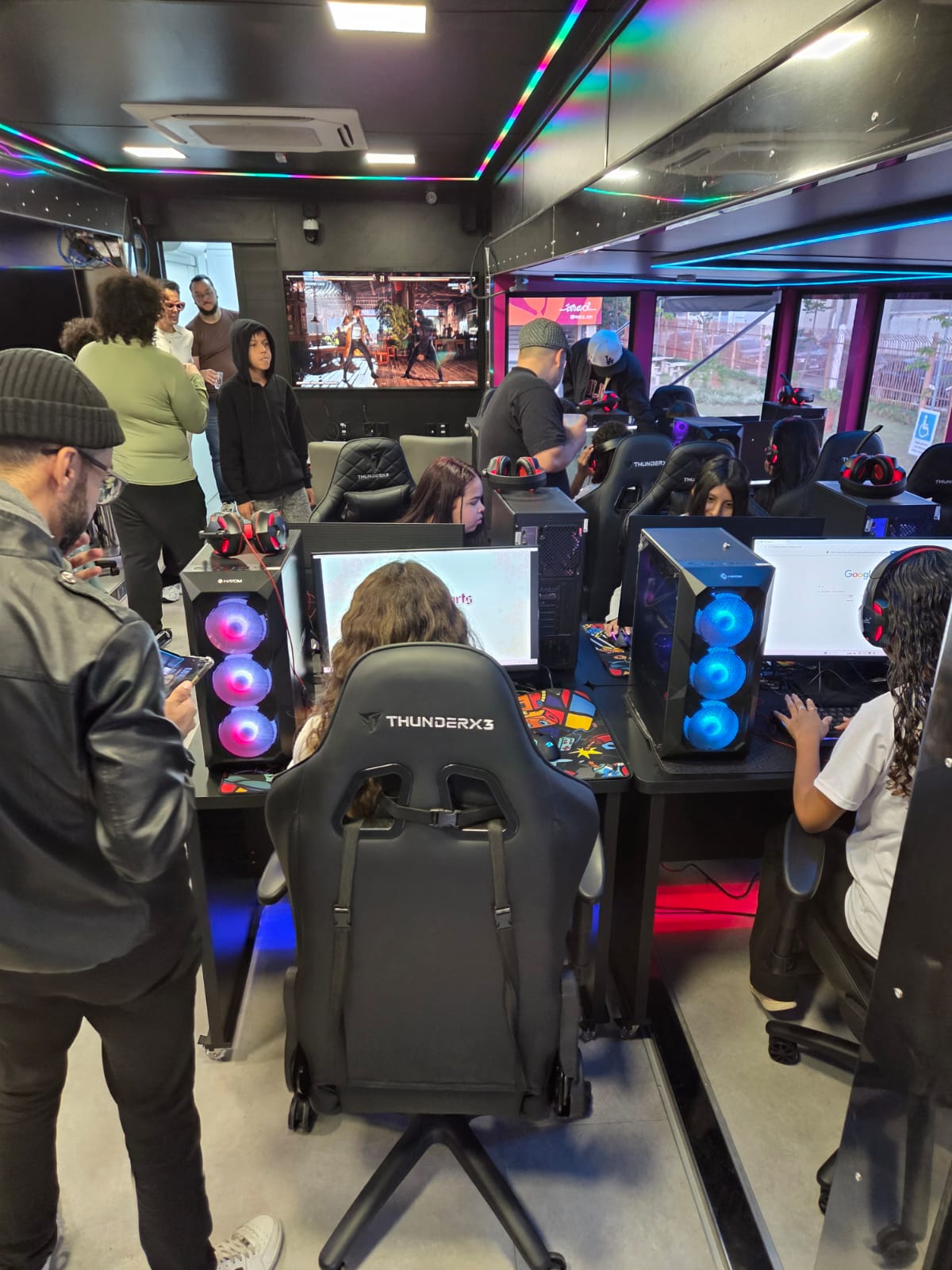 Pessoas utilizam computadores em um espaço interno com estrutura gamer, enquanto outras observam ao redor. O ambiente conta com iluminação colorida, telas com jogos e equipamentos de tecnologia, indicando uma atividade voltada à inclusão digital ou entretenimento.