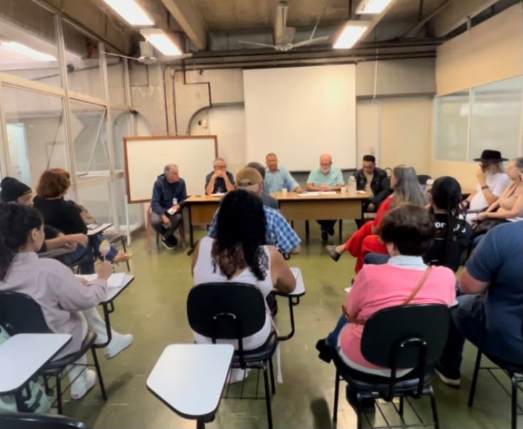 Pessoas participam de uma reunião em uma sala da Subprefeitura de São Miguel Paulista. Os participantes estão sentados em cadeiras organizadas em formato de semicírculo, acompanhando uma mesa composta por representantes que conduzem o encontro. O ambiente conta com quadro branco, projetor e estrutura voltada para discussões e alinhamentos sobre temas de interesse da comunidade e da administração pública.