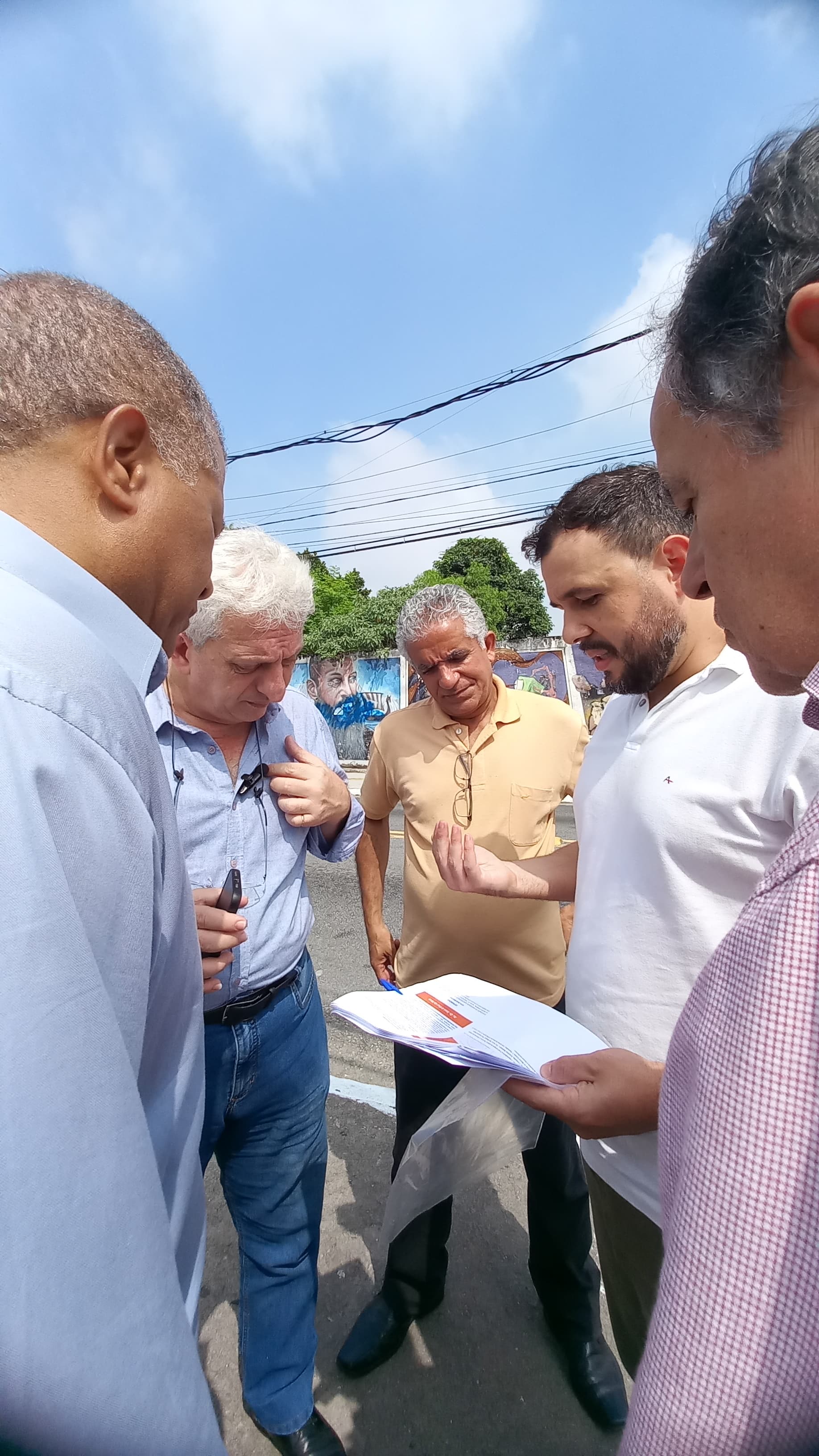 Fotografia mostra cinco homens reunidos ao ar livre, eles estão em pé em um asfalto com um fundo que inclui mural e fios elétricos inclinando-se e examinando um mapa da região.