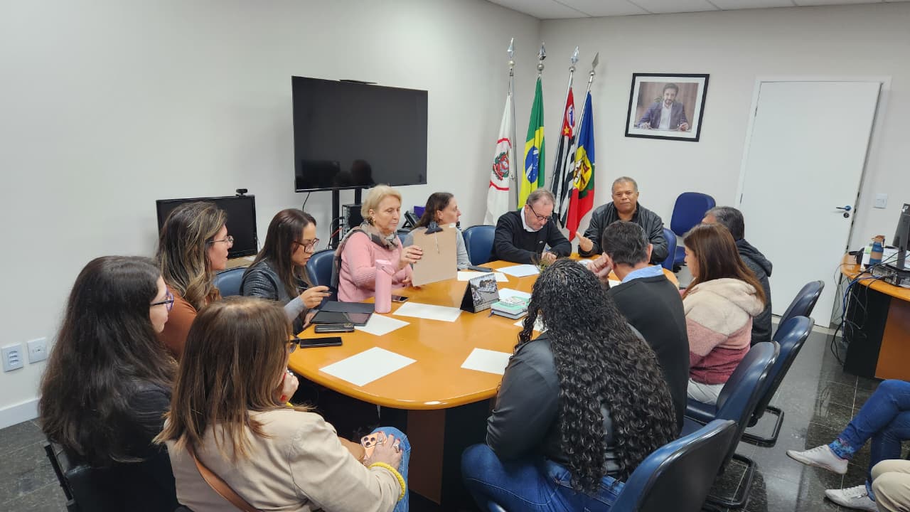Um grupo de pessoas está reunido em volta de uma mesa em uma sala de reunião na Subprefeitura de São Miguel Paulista. Sobre a mesa há documentos e materiais de trabalho sendo analisados. Ao fundo, estão bandeiras oficiais e uma televisão, indicando um ambiente institucional. O encontro registra a reunião que debateu investimentos e melhorias na área da saúde da região.
