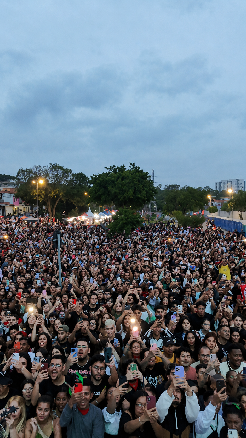 Grande público reunido na Virada Cultural em São Miguel Paulista, em um espaço aberto, com muitas pessoas concentradas e segurando celulares para registrar o evento. Ao fundo, há árvores, tendas, iluminação pública e prédios, indicando a realização de um evento urbano de grande porte.