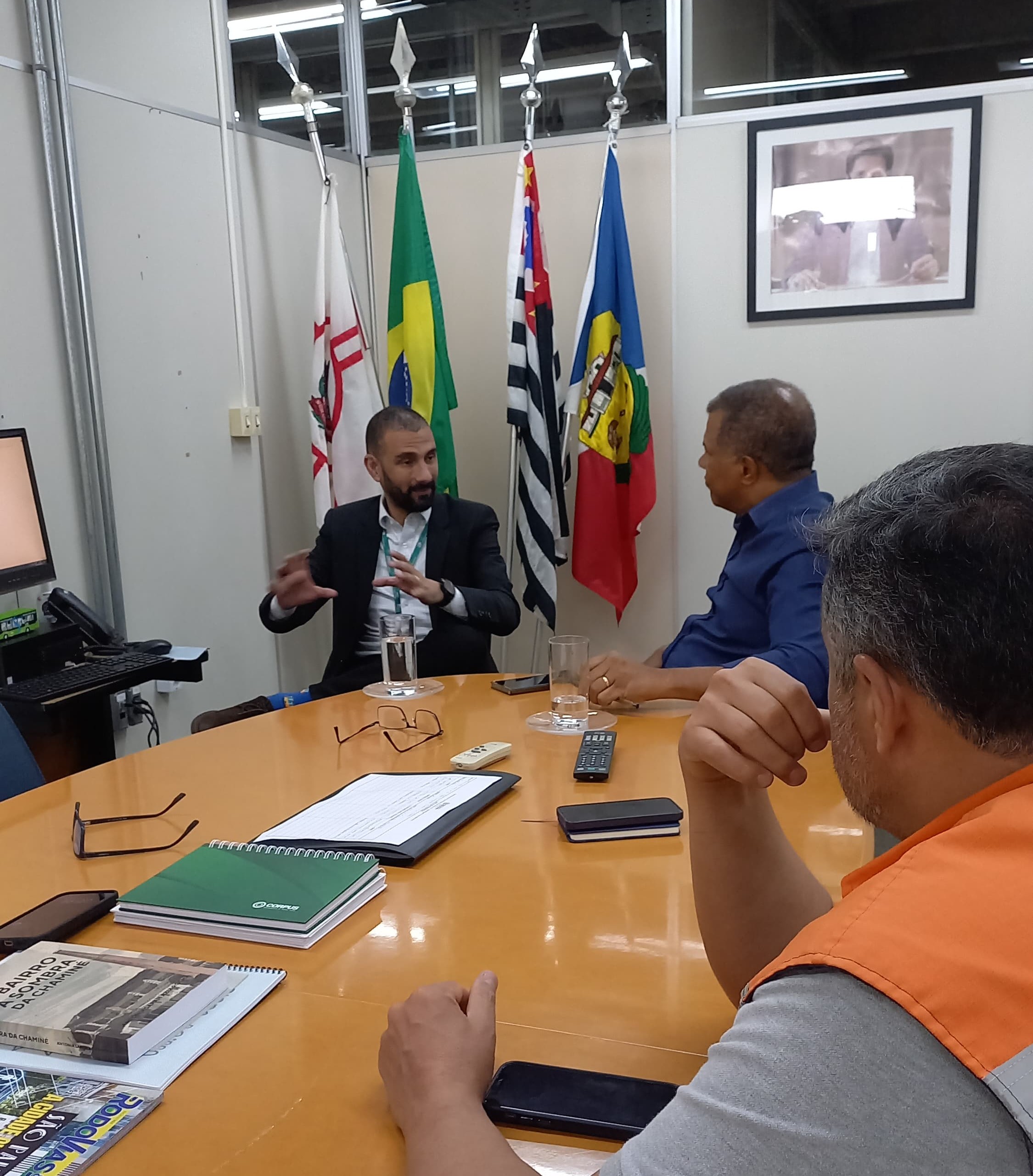 A imagem mostra uma reunião em uma sala de conferências com quatro homens sentados ao redor de uma mesa oval. Um homem em terno está falando, enquanto os outros escutam atentamente. Atrás deles, há três bandeiras e um quadro na parede.