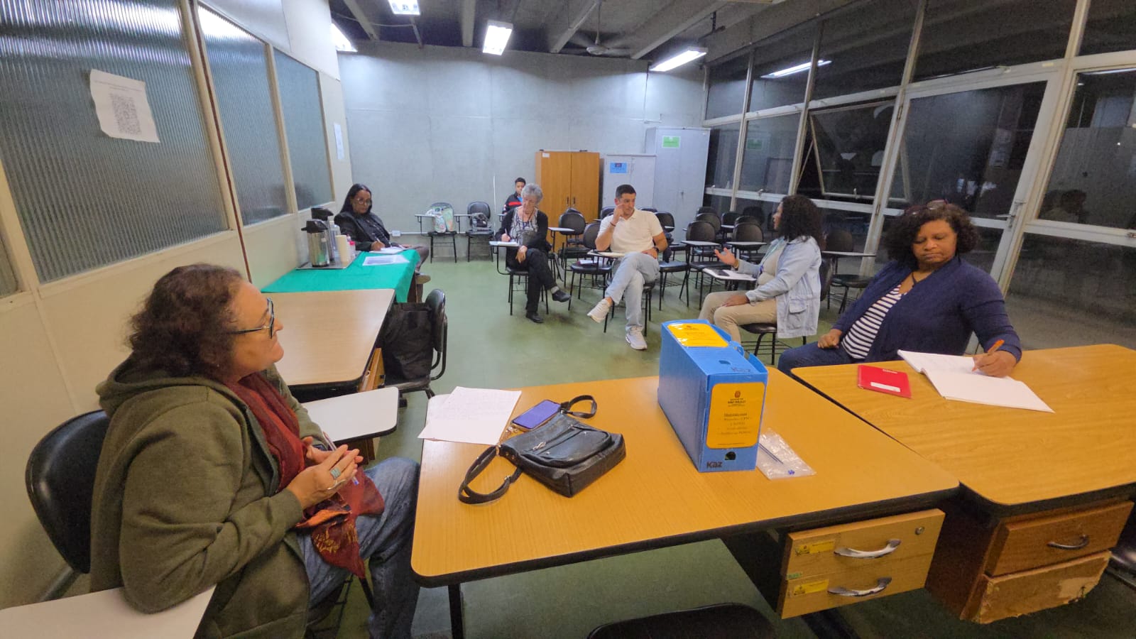 A imagem mostra uma sala de aula ou um ambiente de treinamento com várias pessoas sentadas em mesas individuais, possivelmente participando de uma aula, workshop ou sessão de exame. As pessoas estão sentadas em fileiras, de frente para a parte da frente da sala (fora do campo de visão). A maioria dos participantes parece estar focada em papéis ou anotações em suas mesas. Uma mulher em primeiro plano, à esquerda, usa um casaco verde e um cachecol vermelho.