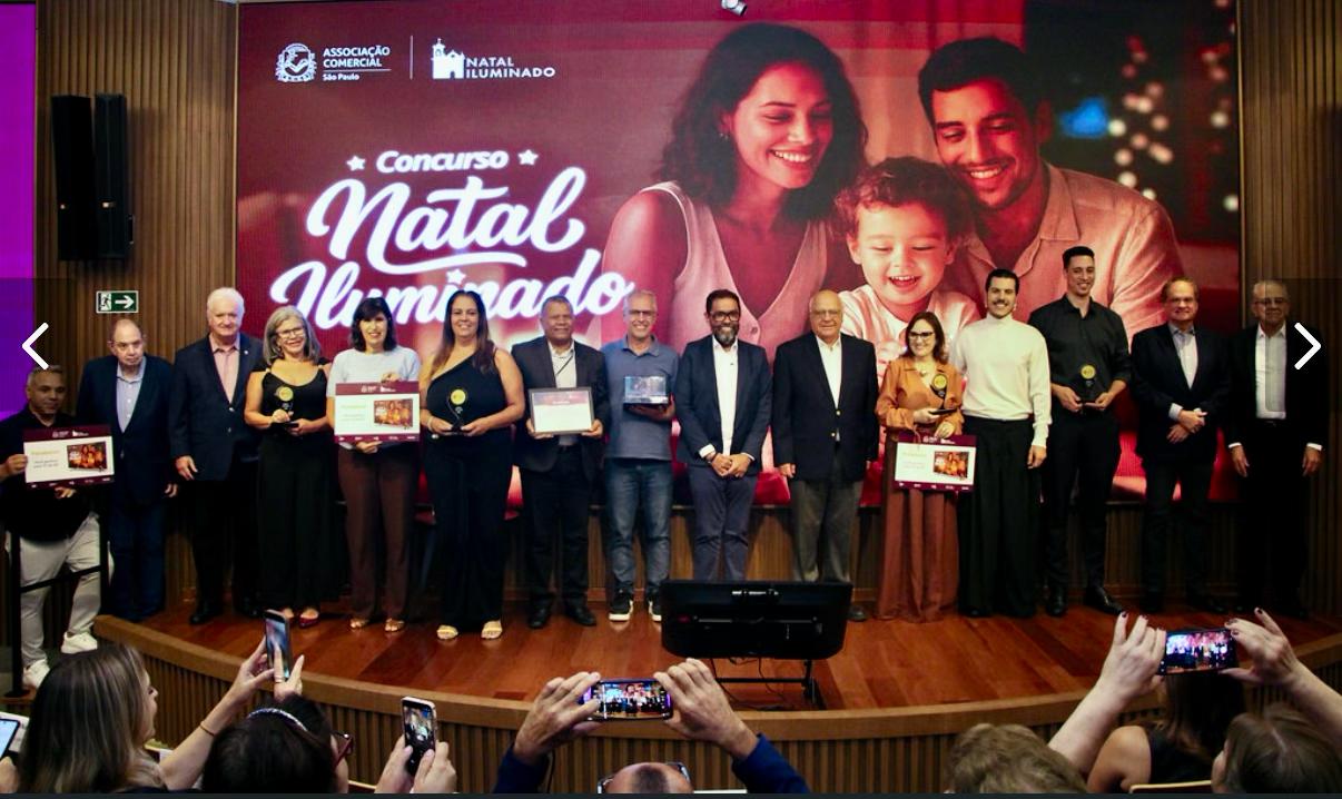 ​A imagem mostra um grupo de 14 pessoas (homens e mulheres) posicionados lado a lado em um palco de madeira, durante uma cerimônia de premiação. Ao fundo, há um grande painel digital vermelho e brilhante. Na parte inferior da foto, em primeiro plano, aparecem mãos da plateia segurando celulares para registrar o momento.