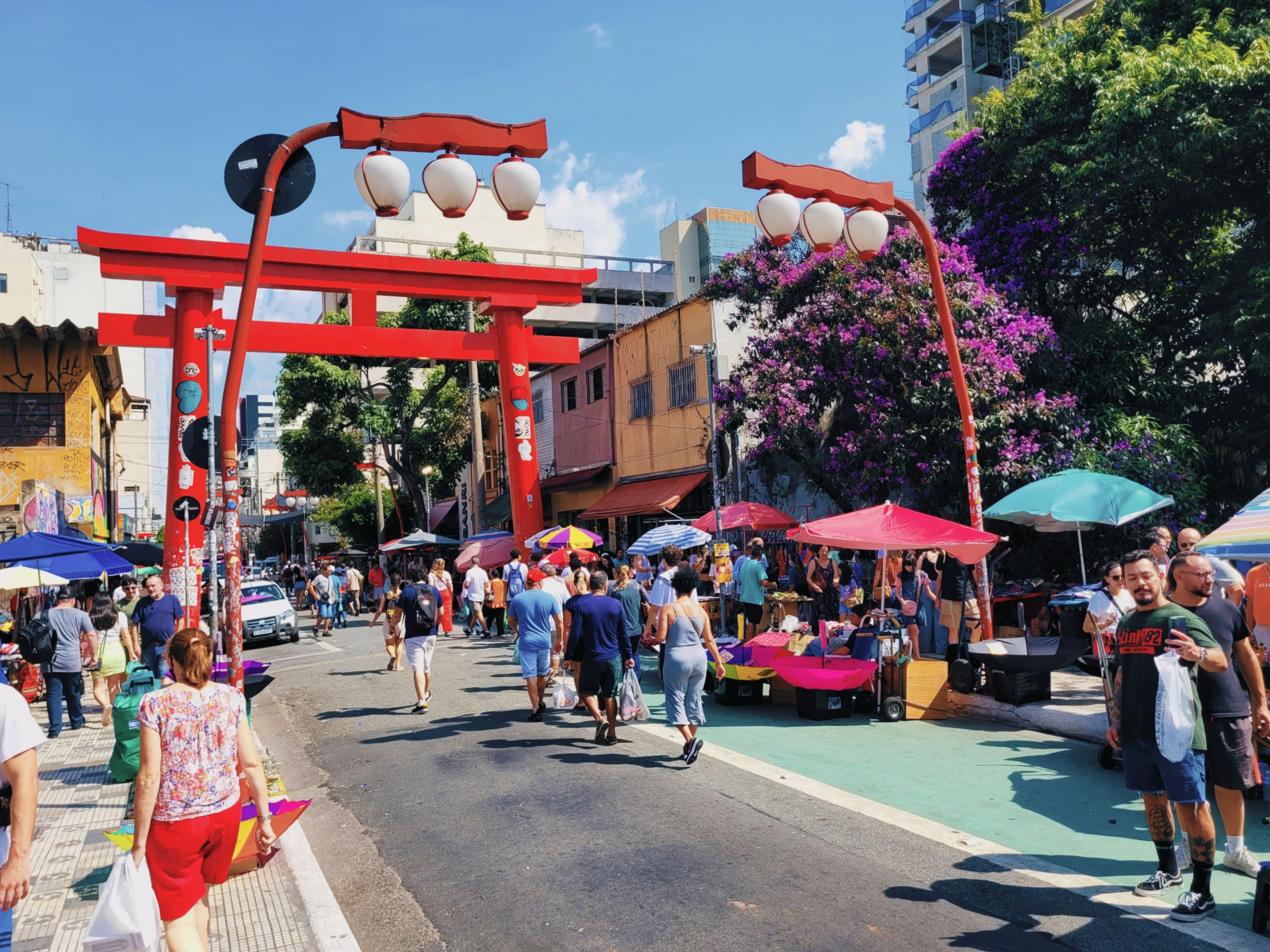 Diversas pessoas caminhando na rua e calçadas da liberdade, algumas vendas de produtos, e alguns guarda-sóis para proteger do sol